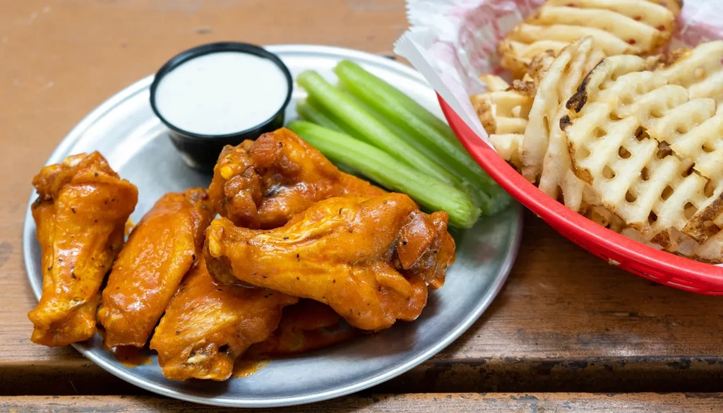 A plate of buffalo wings, celery, dipping sauce, and waffle fries.