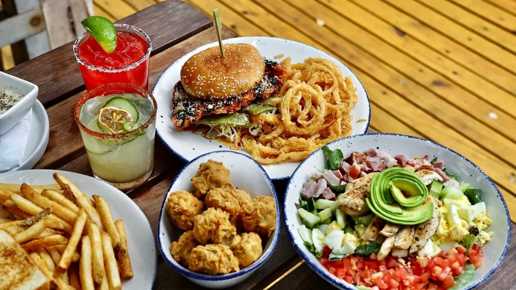 Table with burger, colorful salad, fried chicken, fries, and two cocktails.