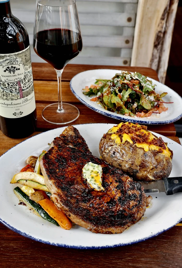 Grilled steak, baked potato, vegetables, salad, and red wine on wooden table.