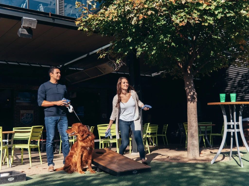Man and woman play cornhole with dog on a restaurant patio.