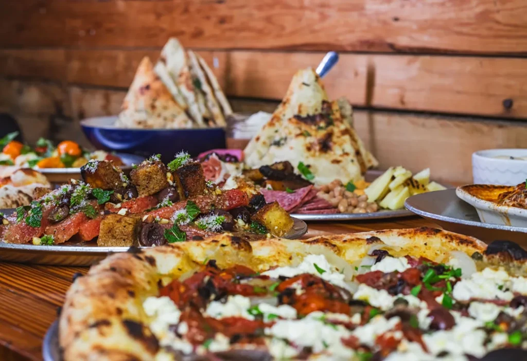 Wooden table with lavish food spread: pizza, salad, charcuterie, flatbread.