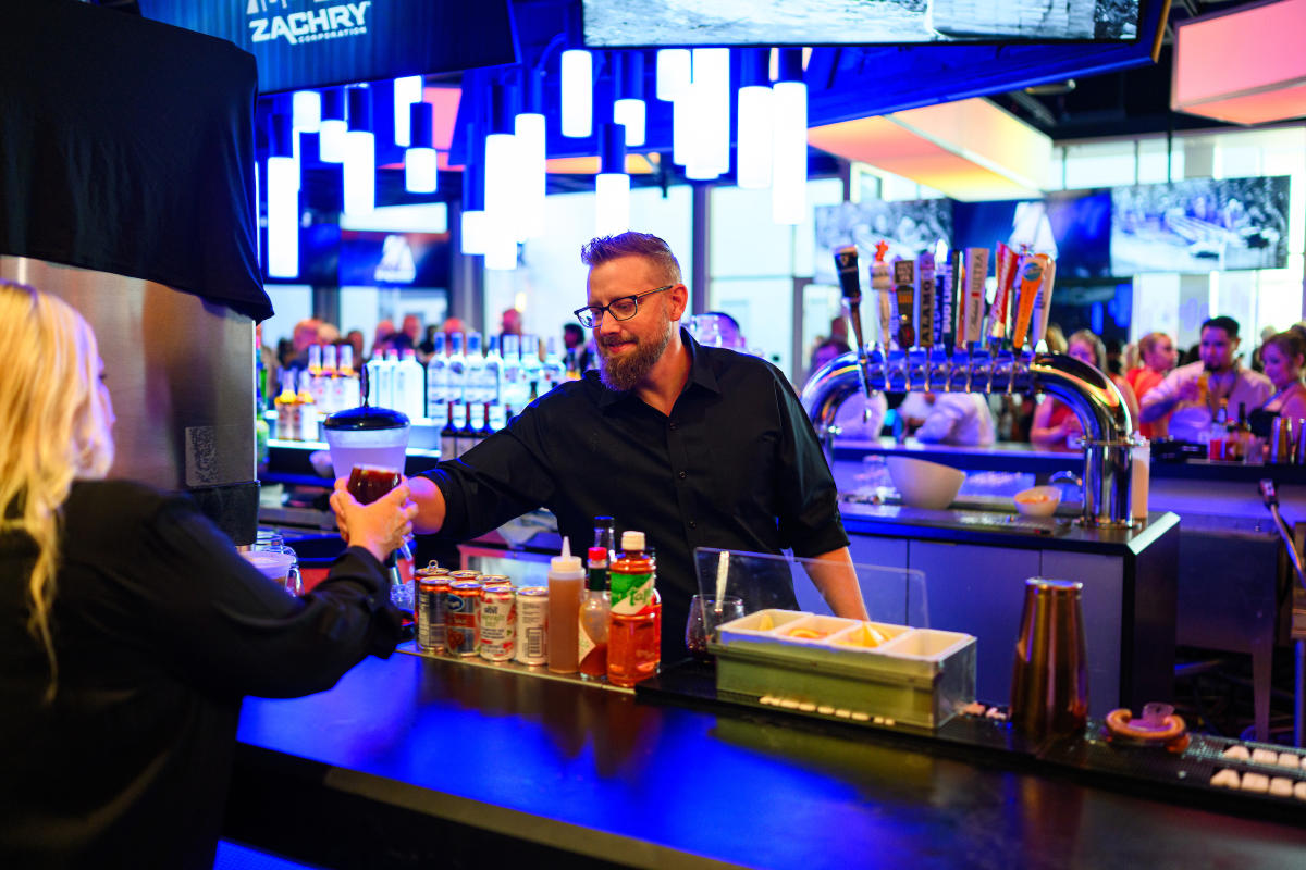 Bartender with beard hands drink to blonde customer at blue-lit bar.