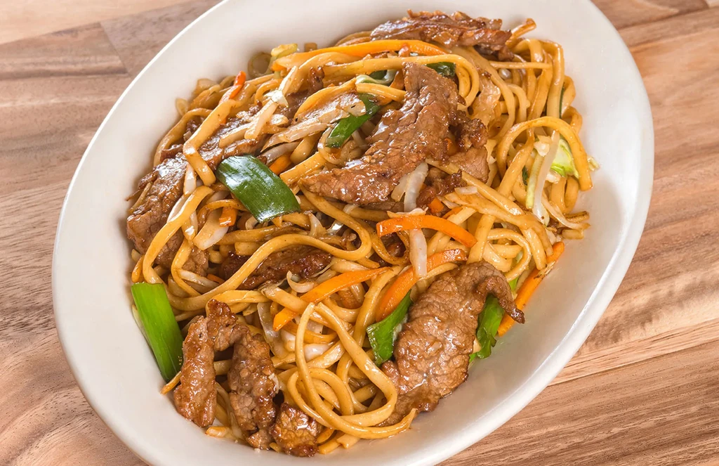 Overhead view of beef chow mein with vegetables on a wooden table.
