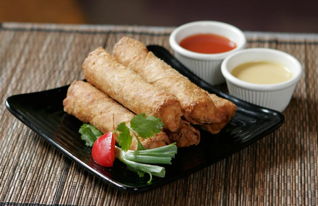 Golden spring rolls on black plate with garnish and sauces.