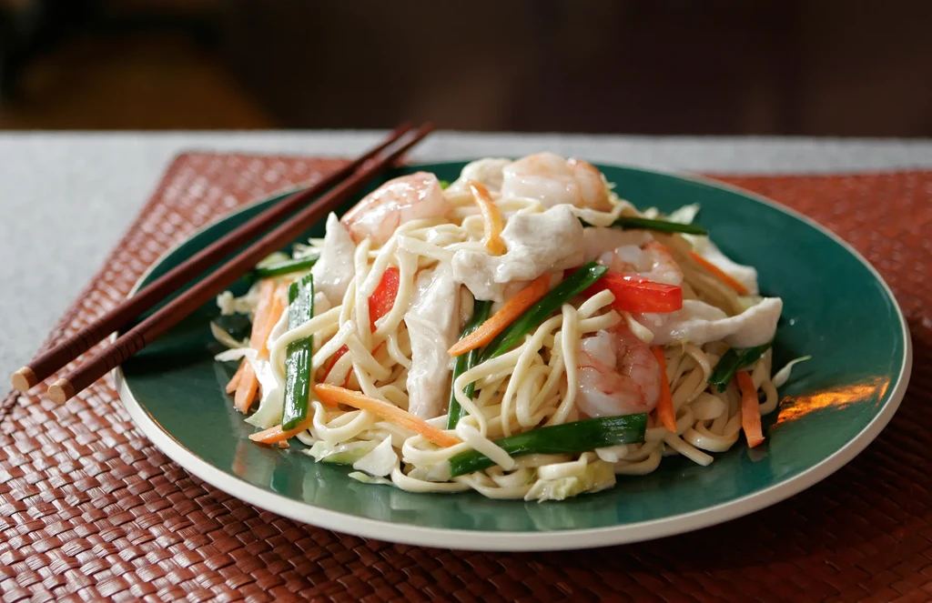 Close-up of stir-fried noodles with shrimp, chicken, vegetables, and chopsticks.