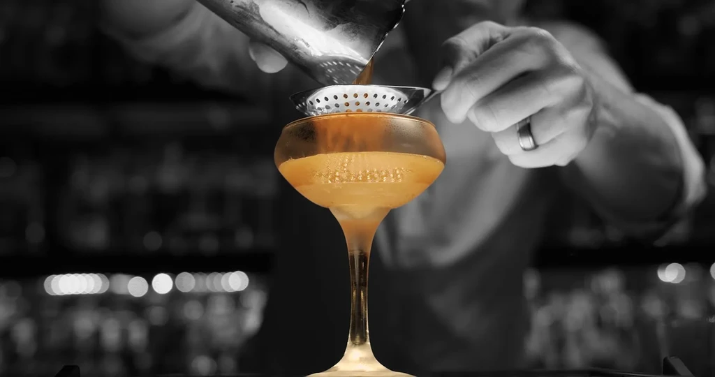 Bartender pours golden cocktail into frosty glass; image partially grayscale.