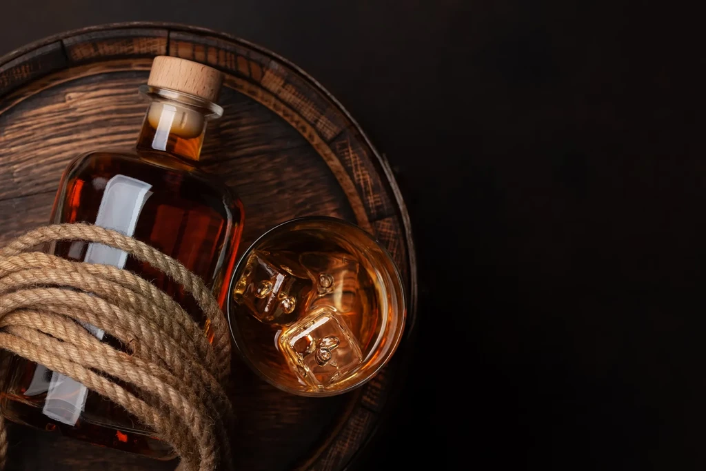 Whiskey bottle, glass with ice, and rope on a dark barrel.