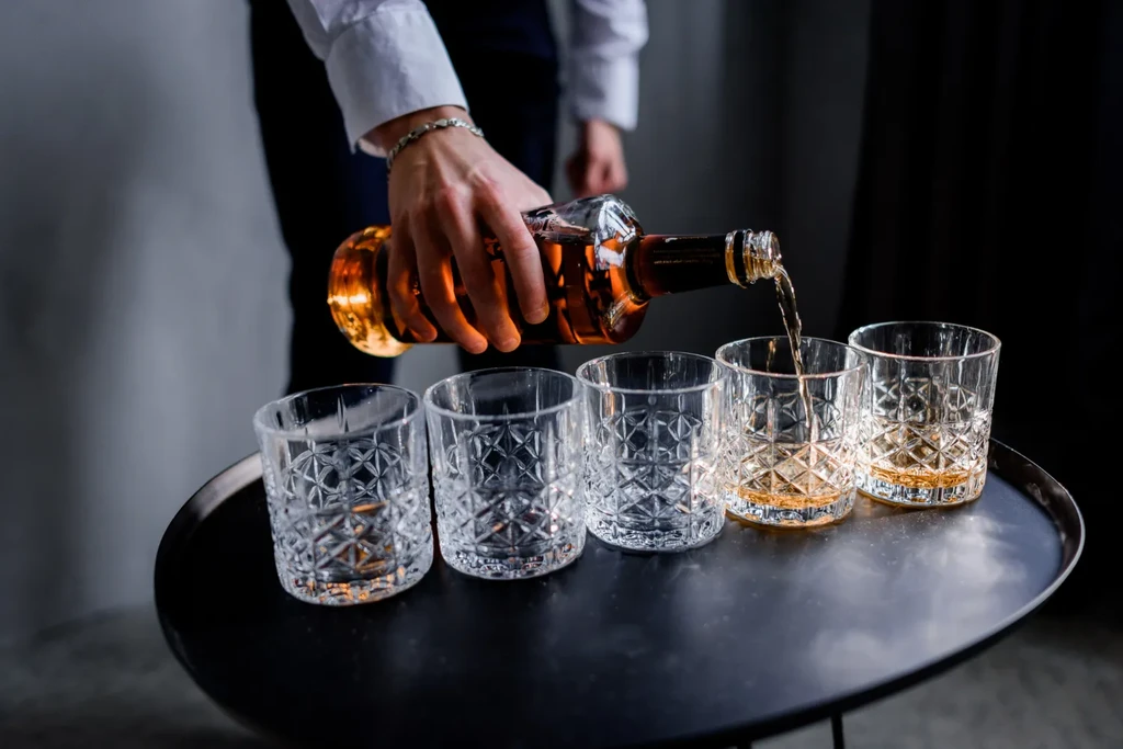Hands pour dark whiskey from a clear bottle into textured tumblers.