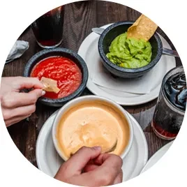 Hands dipping chips into guacamole, salsa, and queso on a table.
