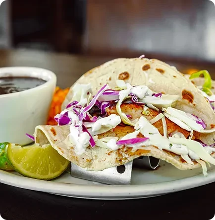 A fish taco with shredded cabbage, creamy sauce, and lime wedge.
