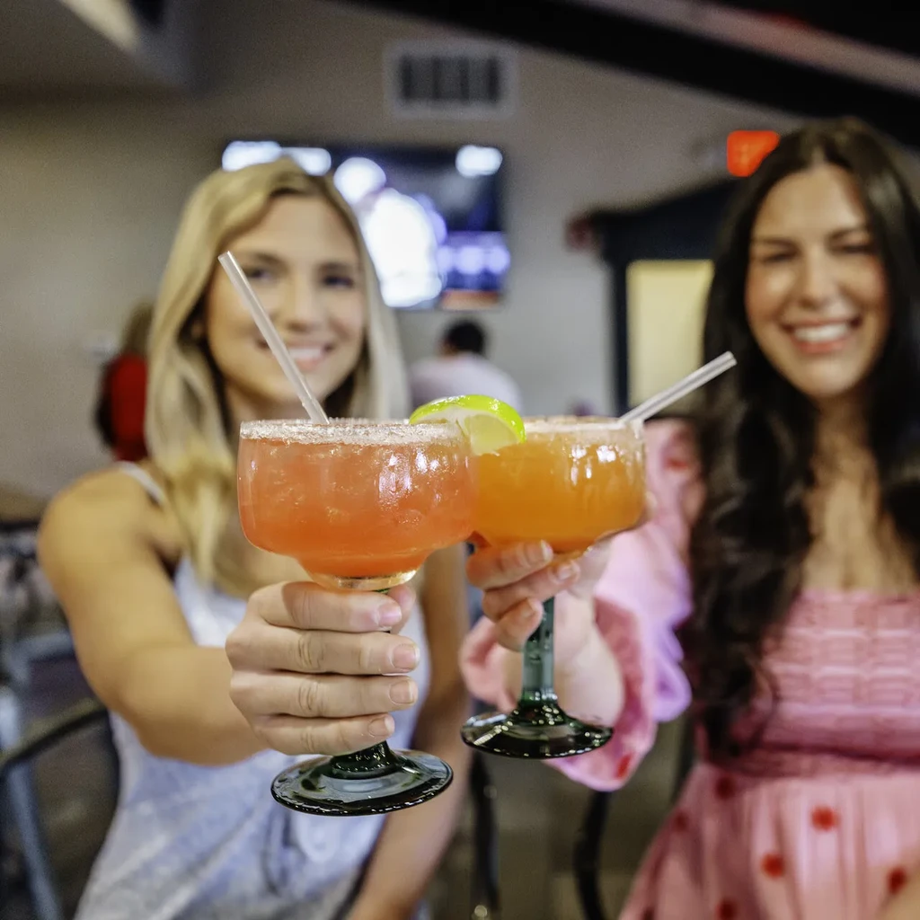 Two vibrant, salt-rimmed cocktails held by people in a dim bar.