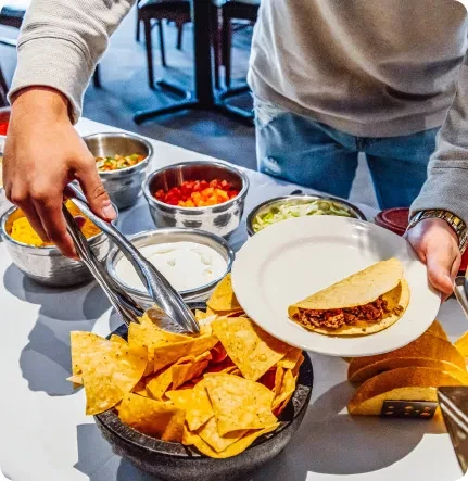 Self-serve taco bar with hands adding chips, cheese, and tomatoes.
