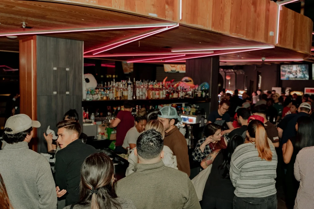Crowded bar at night with people, red neon lights, and bottles.