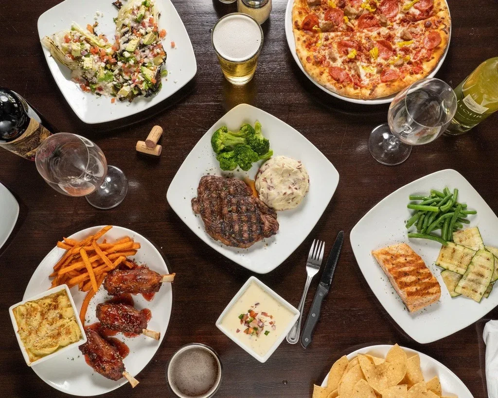 Restaurant table laden with pizza, steak, salmon, sides, and drinks.
