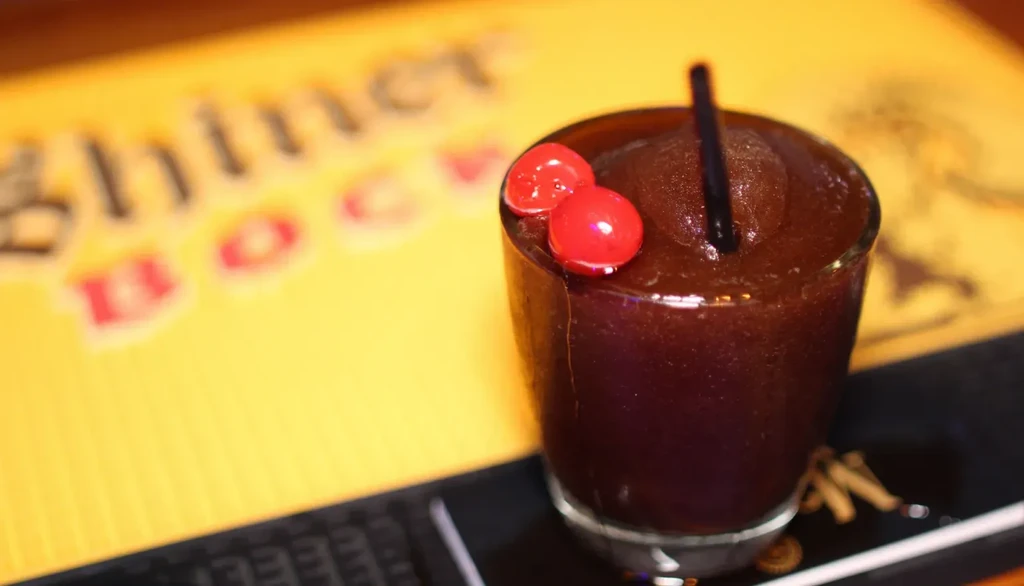 Dark frozen drink with red cherries and black straw in glass.