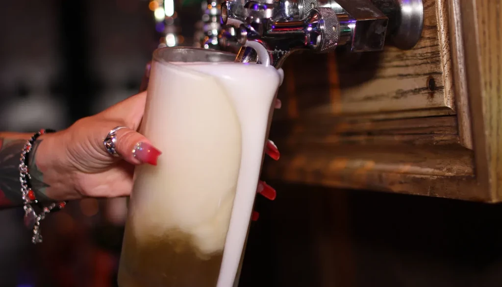 Hand fills beer glass from tap, frothy head overflows.