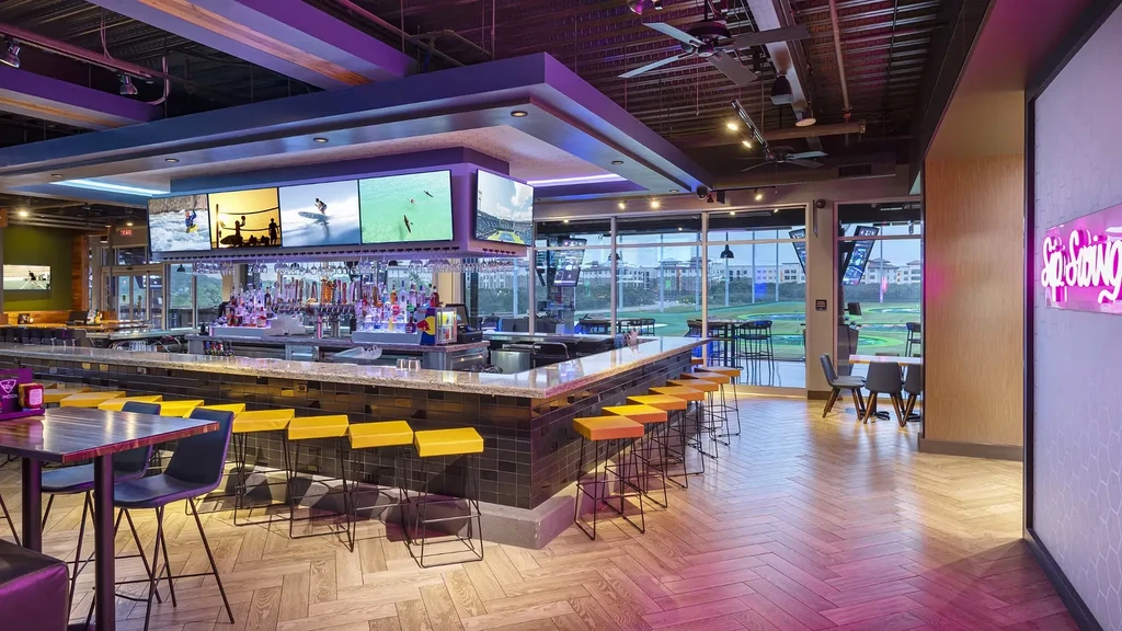 Modern bar with sports TVs, yellow stools, and golf driving range view.