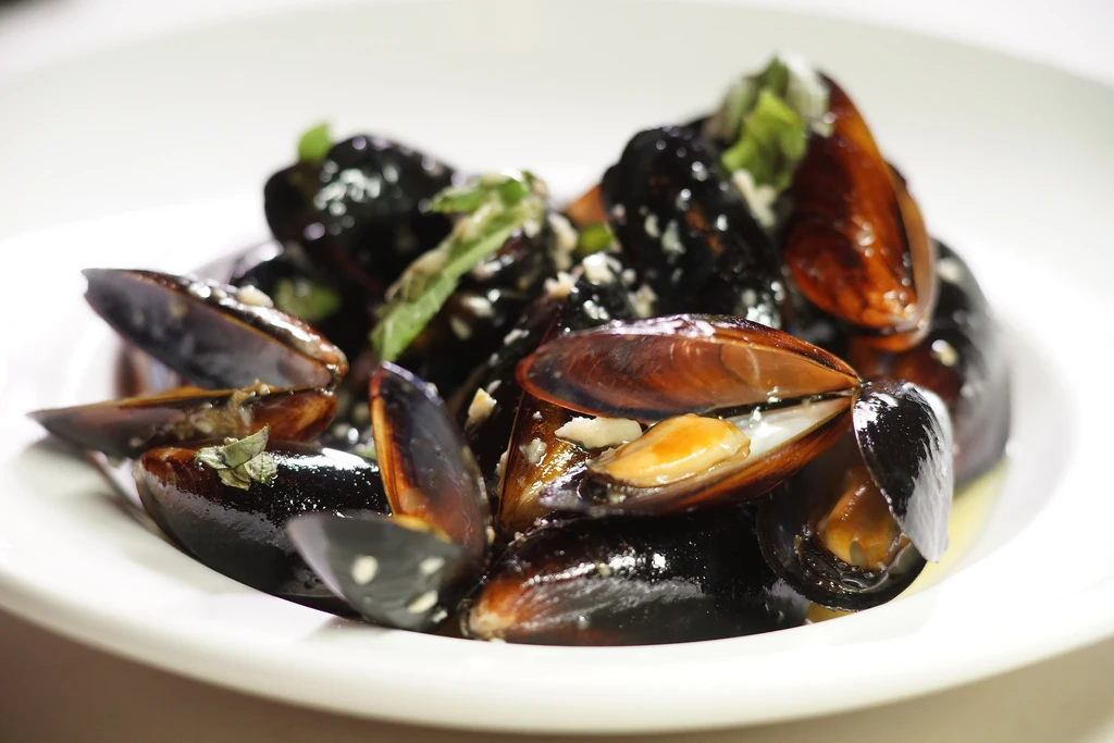 White bowl of steamed mussels, garnished with garlic, herbs, and broth.