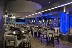 Modern bar interior with tables, a long bar, and blue-lit pillar.
