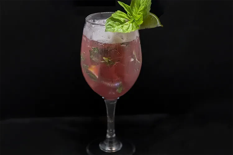Red cocktail in stemmed wine glass with ice, muddled herbs, basil, and lime.