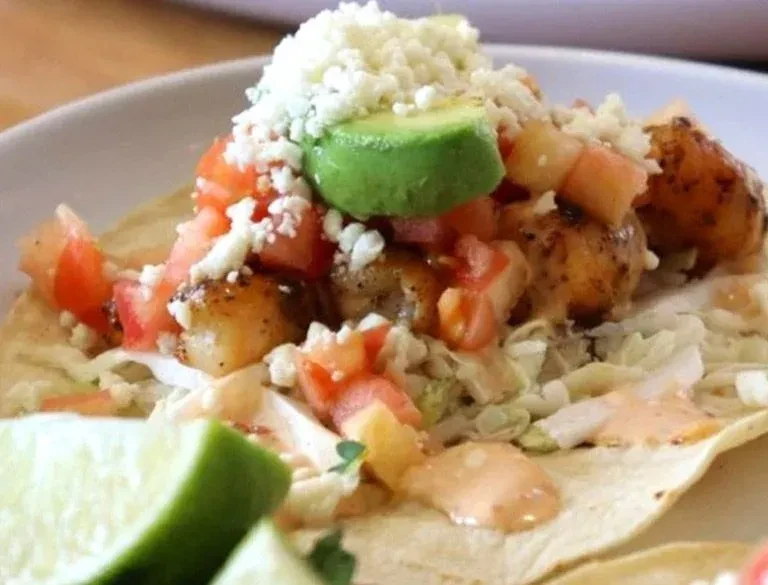Fish or shrimp taco with avocado, lettuce, cheese, and lime wedges.