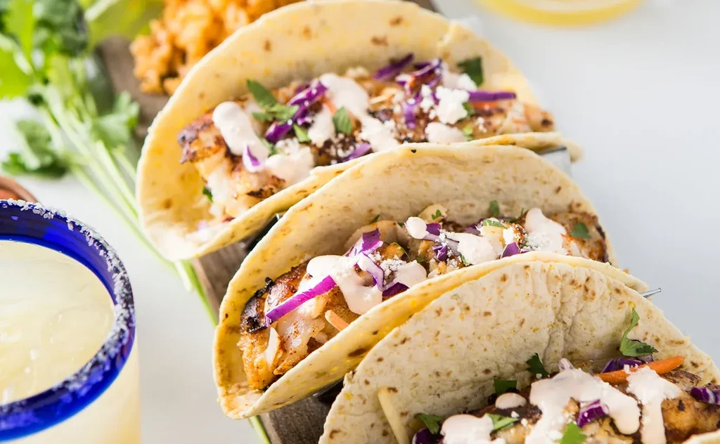 Three fish tacos with purple cabbage, next to a blue-rimmed margarita.