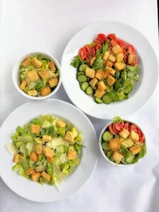 Four salads in white plates and bowls: two Caesar, two mixed greens.