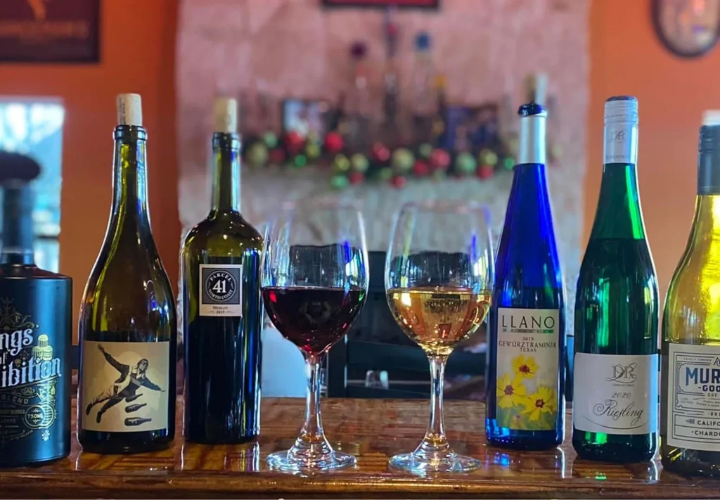Six colorful wine bottles and two filled wine glasses on a wooden bar.