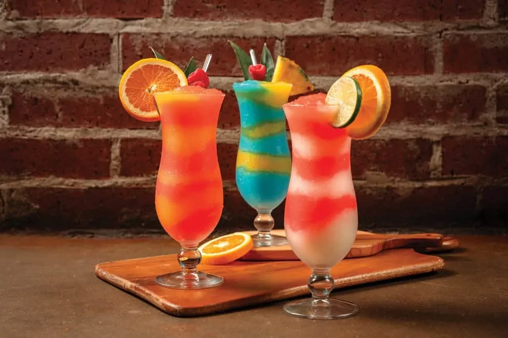 Three colorful frozen drinks on a wooden board against a brick wall.