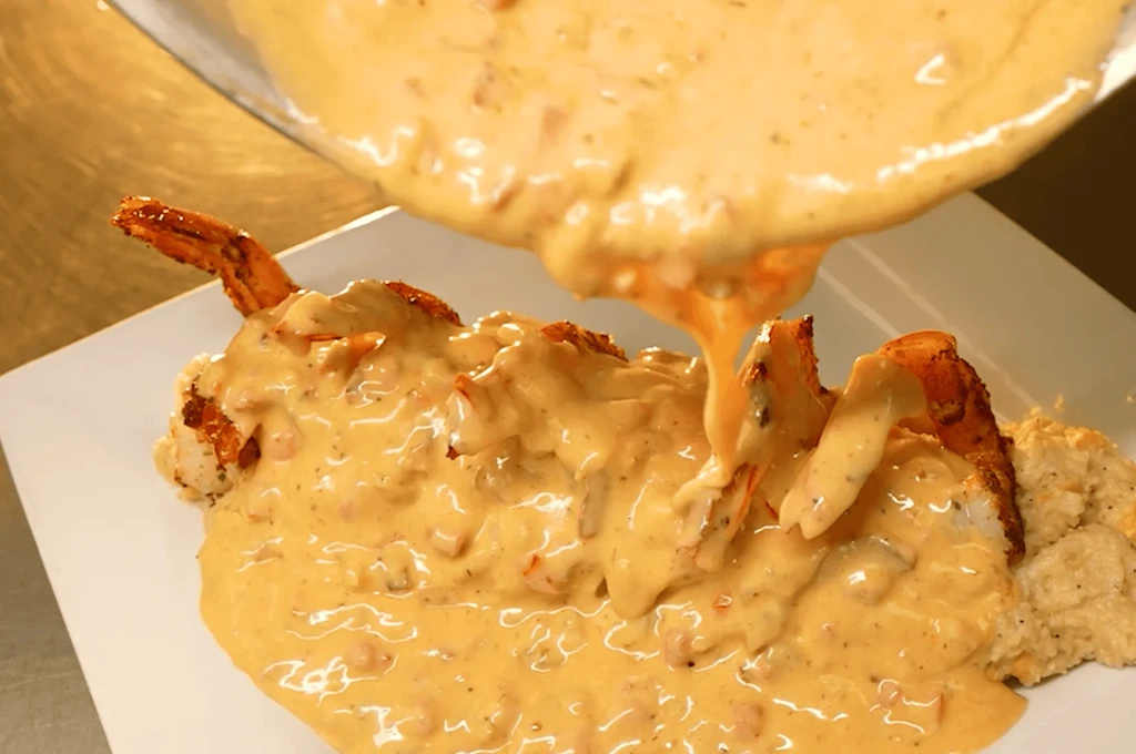 Close-up: Large shrimp and creamy grits being sauced with creamy orange sauce.
