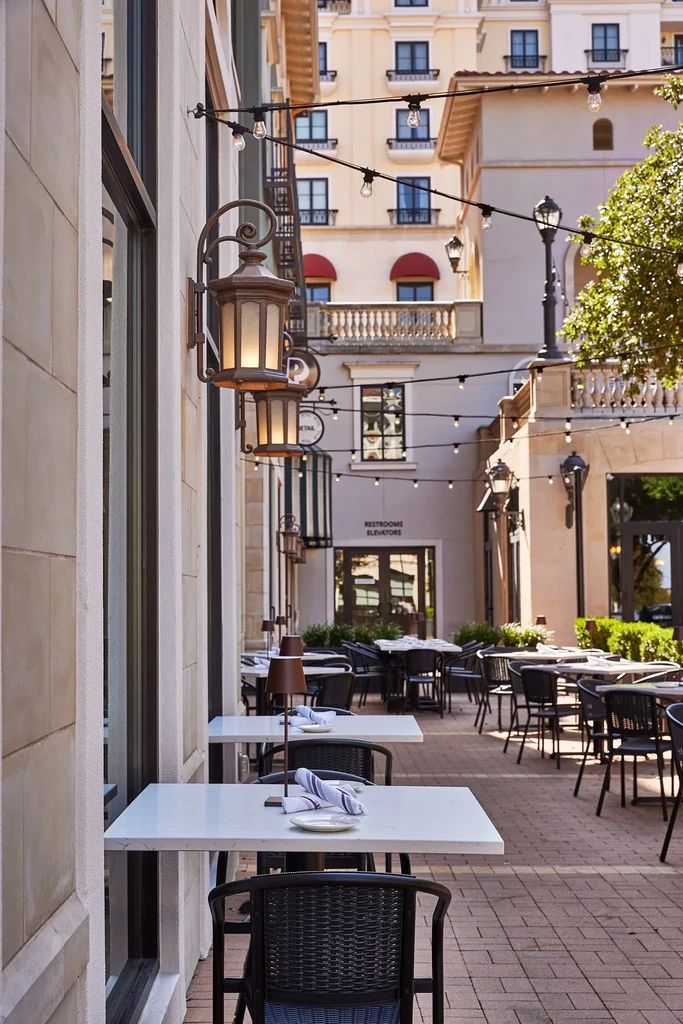 Urban outdoor restaurant patio with set white tables and warm lighting.