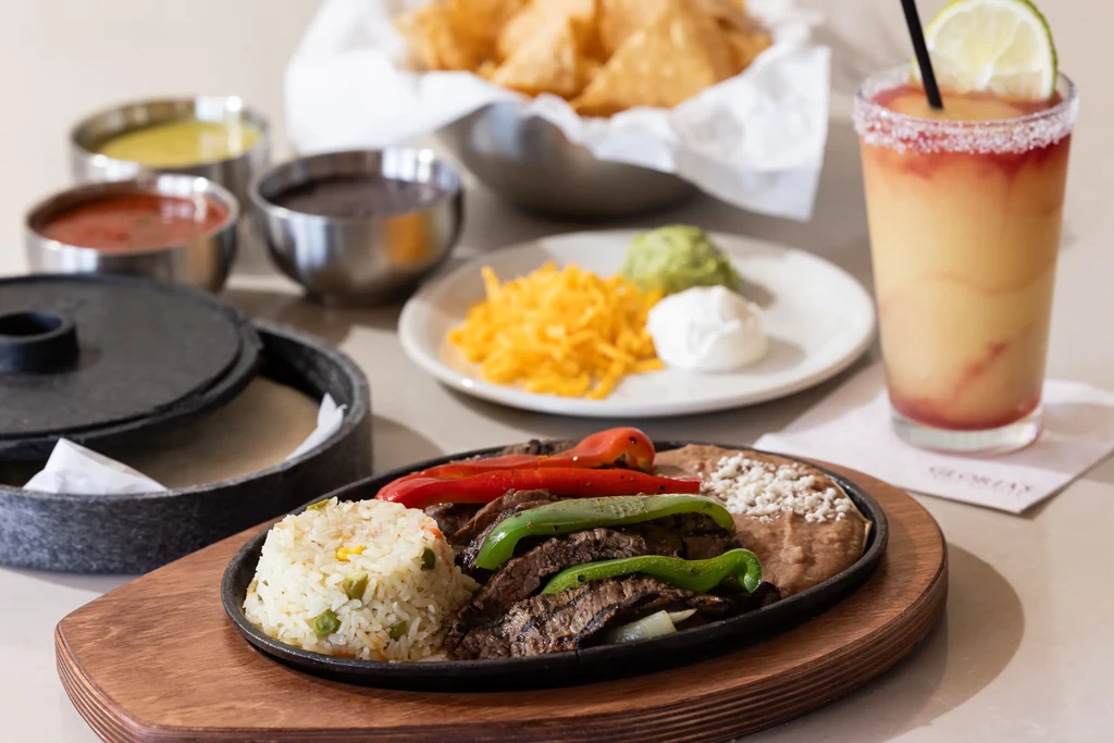 A Mexican food spread with sizzling fajitas, rice, beans, and a drink.