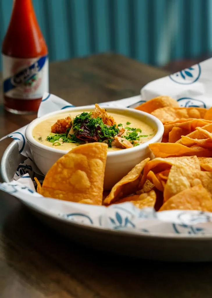 Garnished creamy cheese dip with crispy chips, hot sauce, teal wall.