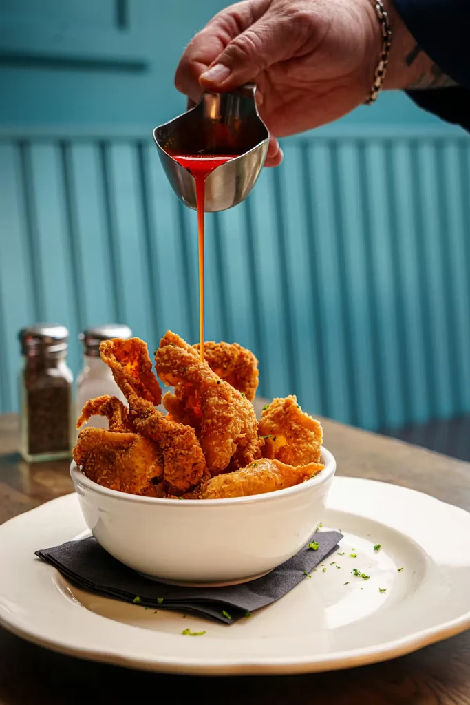 A hand pours red sauce onto crispy fried food in a bowl.