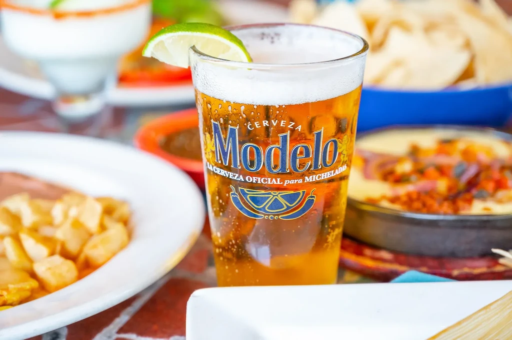 A Modelo beer pint with lime, condensation, and blurred food background.