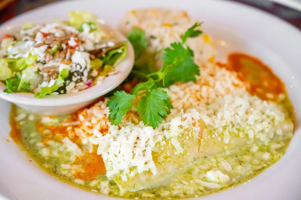Close-up of enchiladas with dual sauces, crumbled cheese, cilantro, side salad.