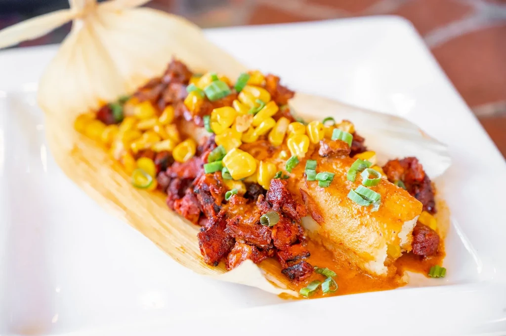 Opened tamale in corn husk with meat, corn, onions, and red sauce.