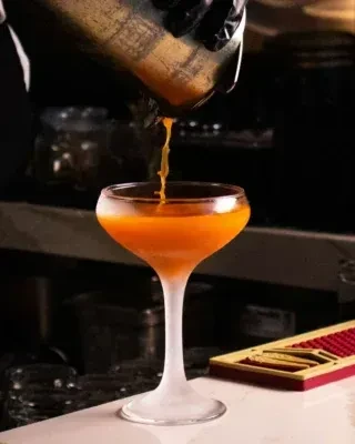 Gloved hand pours vibrant orange cocktail into frosted coupe glass.