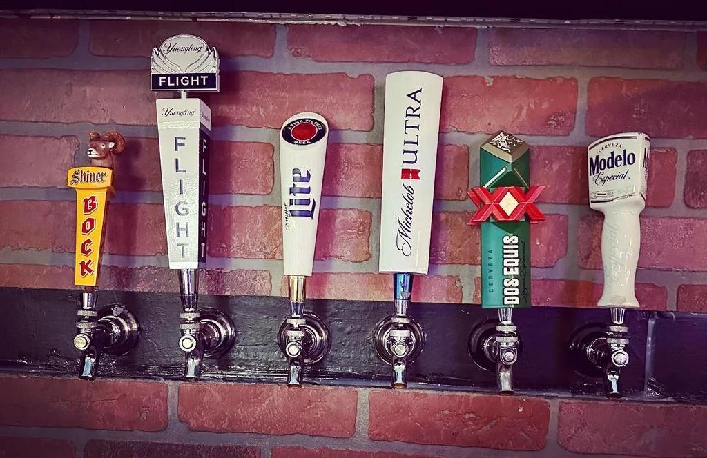 Six branded beer tap handles on a dark board, brick wall.