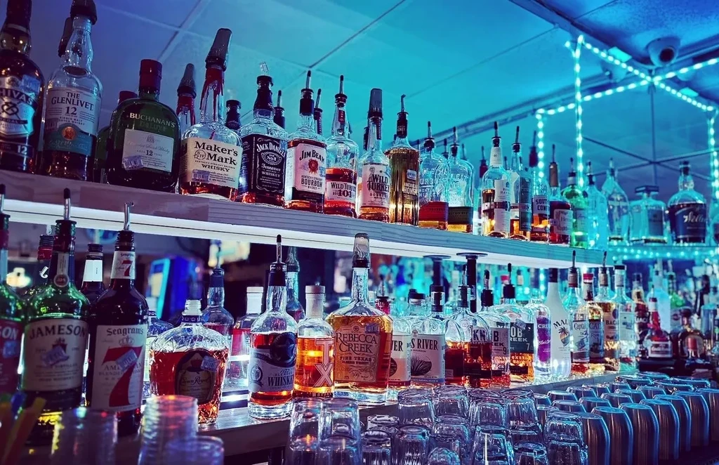 Blue-lit bar displays numerous whiskey bottles, glasses, and cocktail shakers.