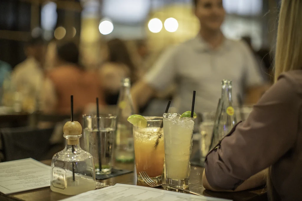Cocktails and drinks on wooden table, blurred restaurant patrons behind.