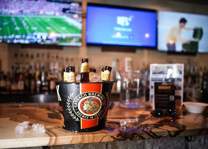 Black Karbach ice bucket, Shiner Bock, on a bar counter with TVs.