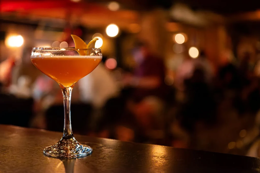 Orange cocktail with foam, citrus peel, blurred bar background.