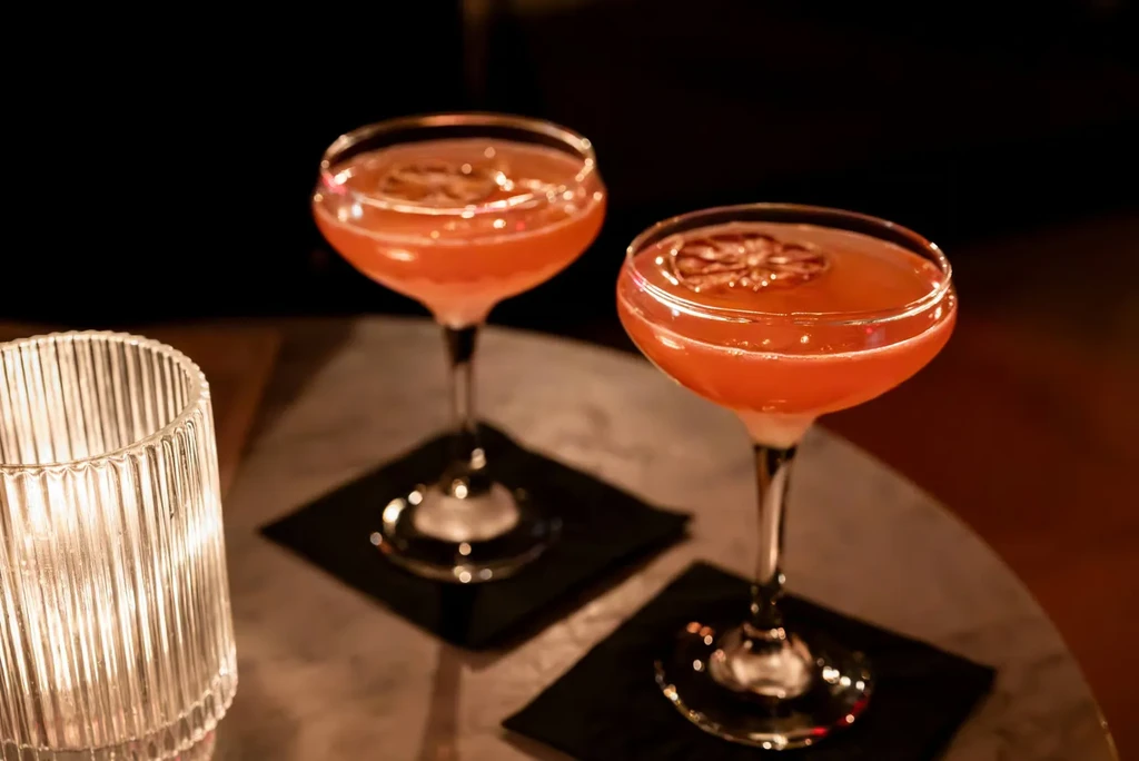 Two reddish cocktails in coupe glasses with citrus, by a glowing candle.