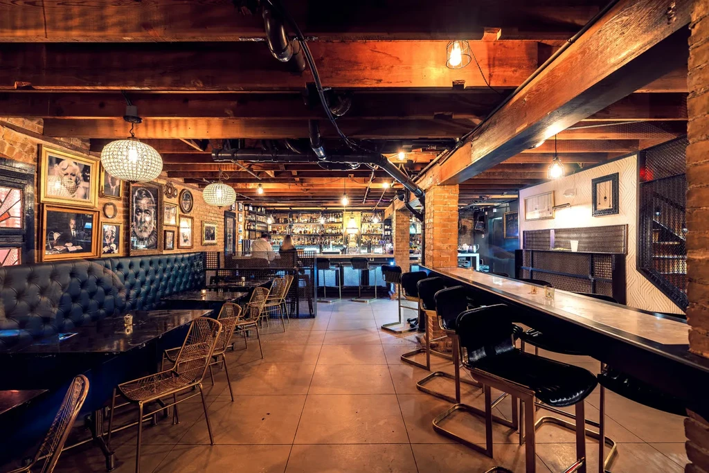 Bar interior with exposed beams, brick walls, tufted booth, and portraits.