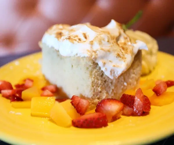 Cake slice with toasted meringue, strawberries, mango, and mint on yellow plate.