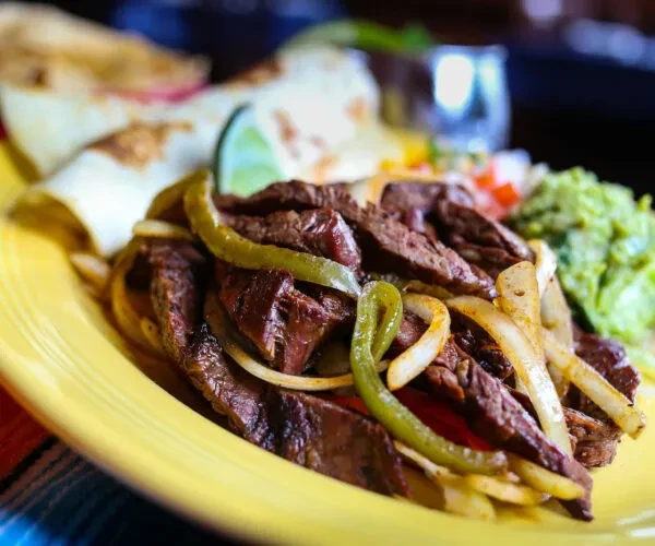 Steak fajitas with peppers, onions, guacamole, salsa, lime, and tortillas.