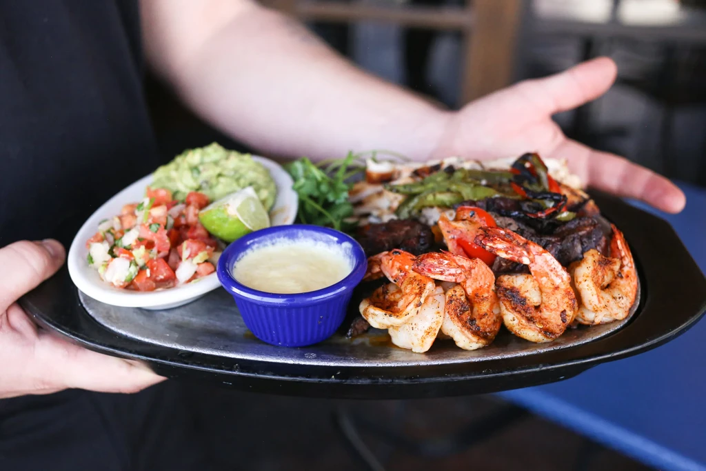 Person carrying sizzling fajita platter with shrimp, meat, vegetables, and dips.