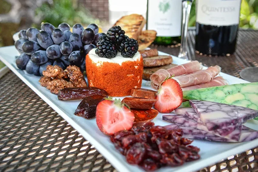 Outdoor charcuterie platter with cheeses, fruits, meats, and wine bottles.