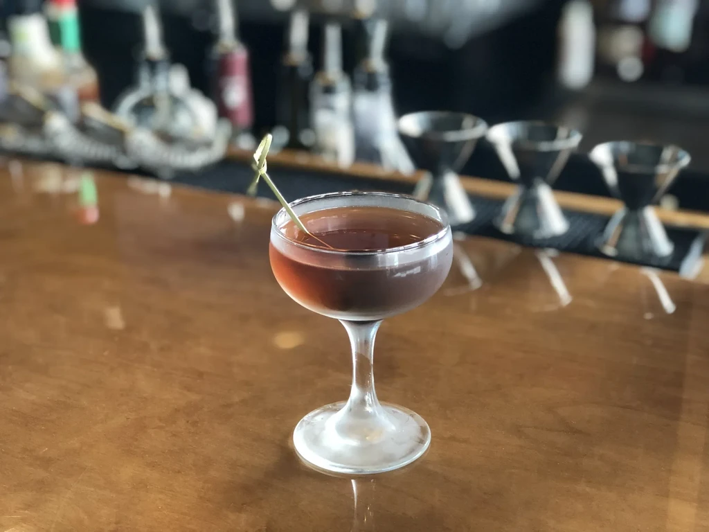 Amber cocktail in a coupe glass with green garnish on bar.
